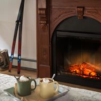 two mugs of hot cocoa on a tray on a bed with a fireplace in thw background with orange flats and a pair of cross country skis leaning against a white wall.