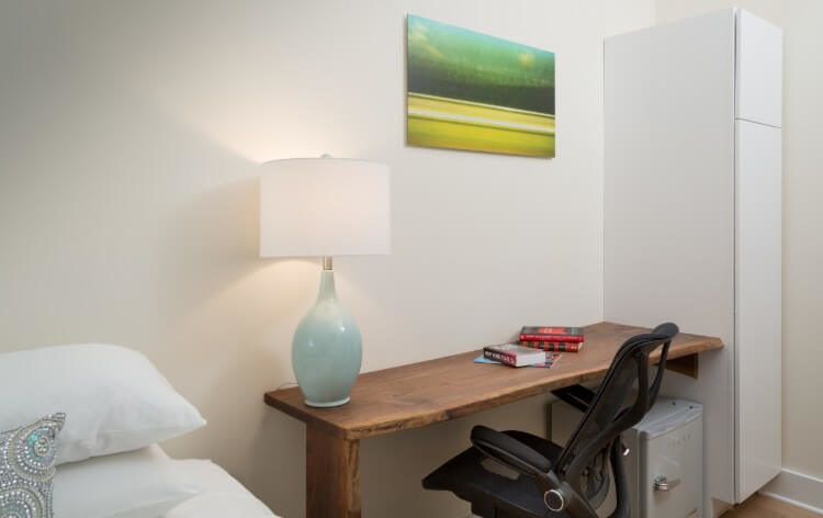 A smooth brown wooden desk with a lamp on the right and a green picture overhead. A black chair is by the desk.