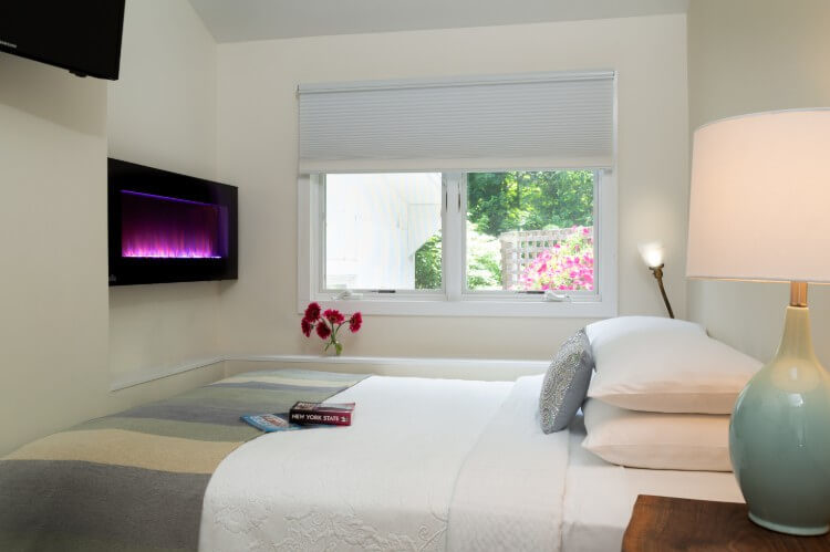 A bed with pillows, a window looking out over green trees and pink flowers, and an electric fireplace on the wall.