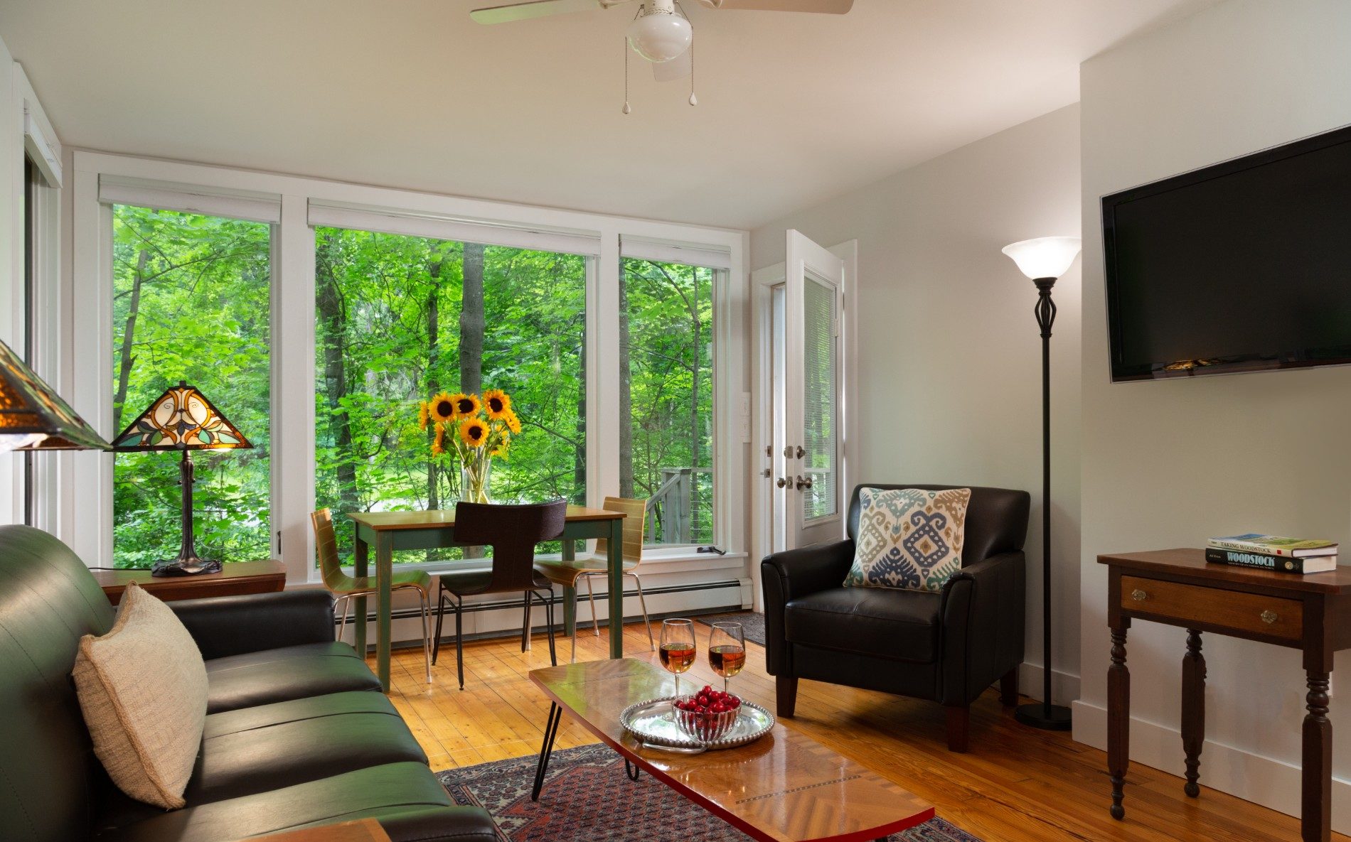 A wooden floor, brown couch, coffee table and flowers on a small table, beside a big floor to ceiling window.