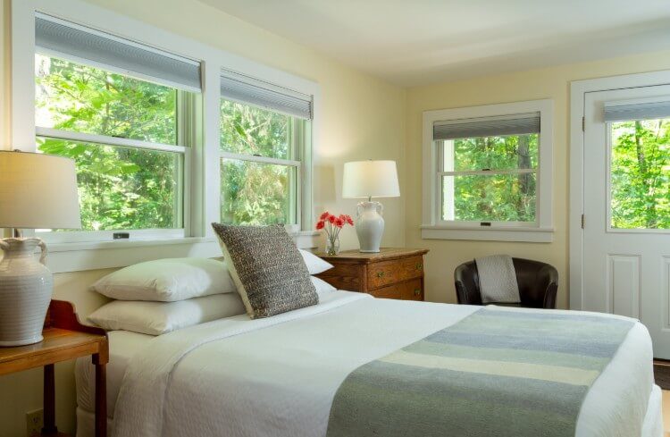 A white bed, with pillows and a lamp on a desk with windows throughout.