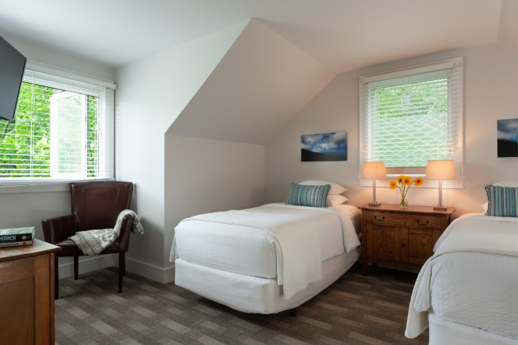 Brown carpet, a twin bed under a dormer ceiling and another twin in the corner. Theres a dresser between the beds and a chair by the window.