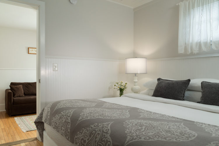 Big bed with white coverlet and gray blanket, overlooking another room with a brown couch.
