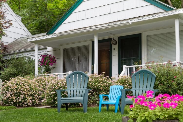 Three Adirondack chairs on green grass, in front of a small cottage with two doors.