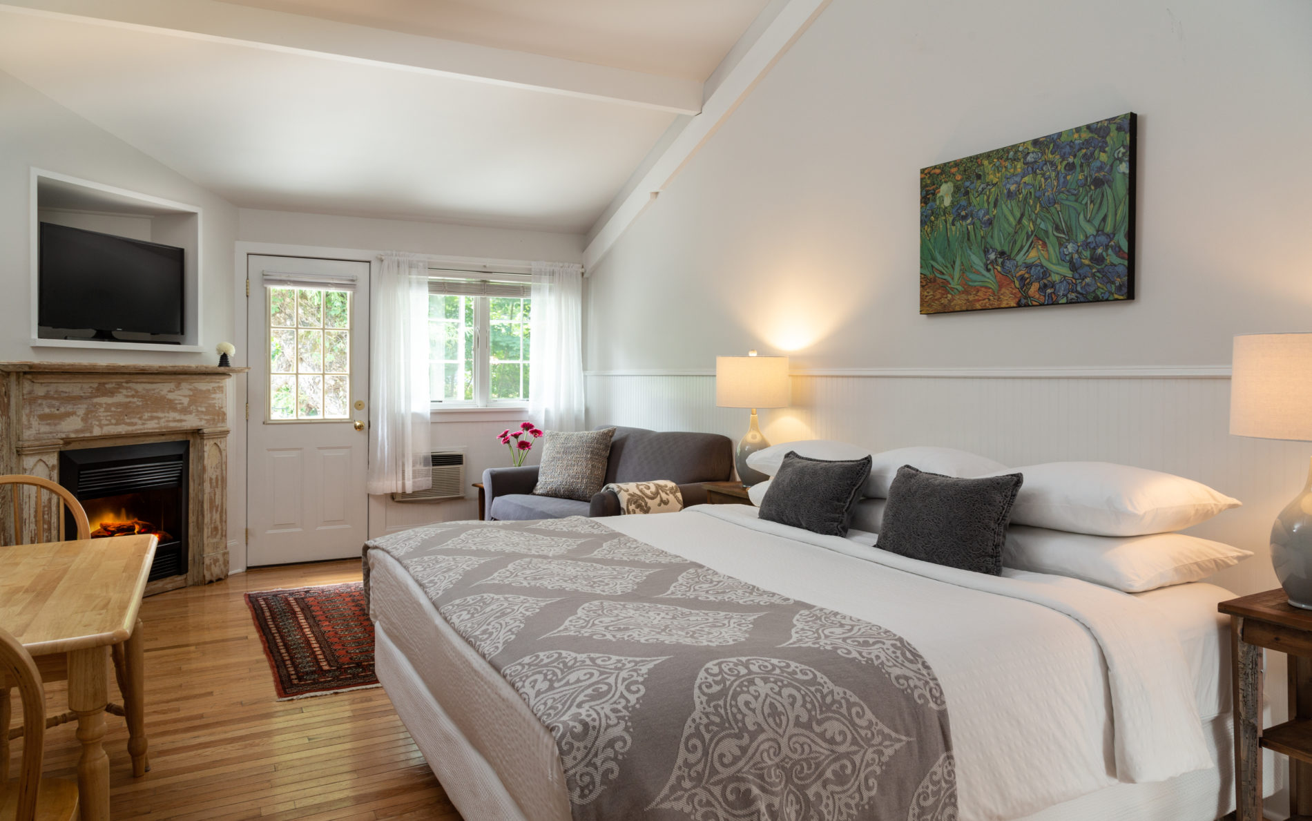 Room with wooden floor, door and big window with sun streaming in, white bed with gray coverlet and a distressed fireplace and small table.