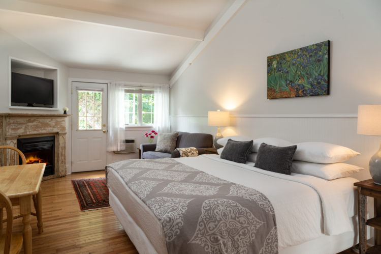 Room with wooden floor, door and big window with sun streaming in, white bed with gray coverlet and a distressed fireplace and small table.