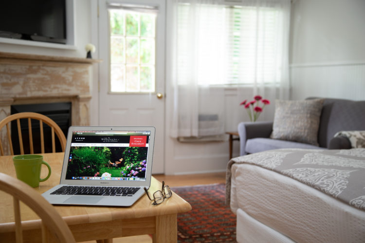 Desk with a computer and cup of coffee next to it, couch, bed and fireplace in the background.