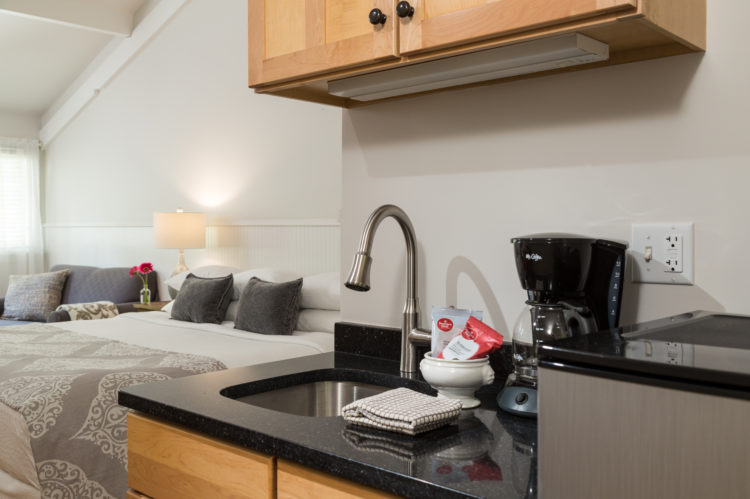 Black counter top with a sink, coffee pot, wooden shelves. In background a white bed with a gray blanket.