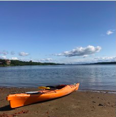 An orange kayak sits on brown sand, by blue water with a few whoite clouds in the sky. 