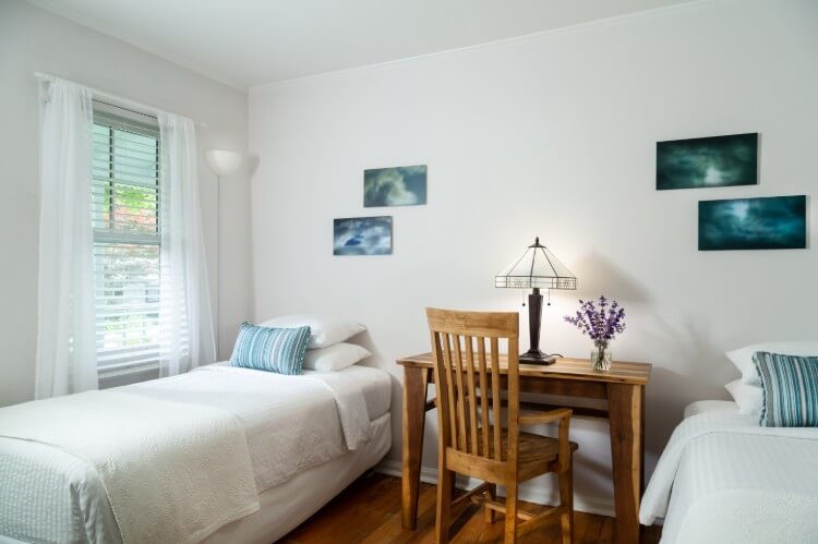 A white room with two twin beds and a desk between them. A large window has sun shining through it.