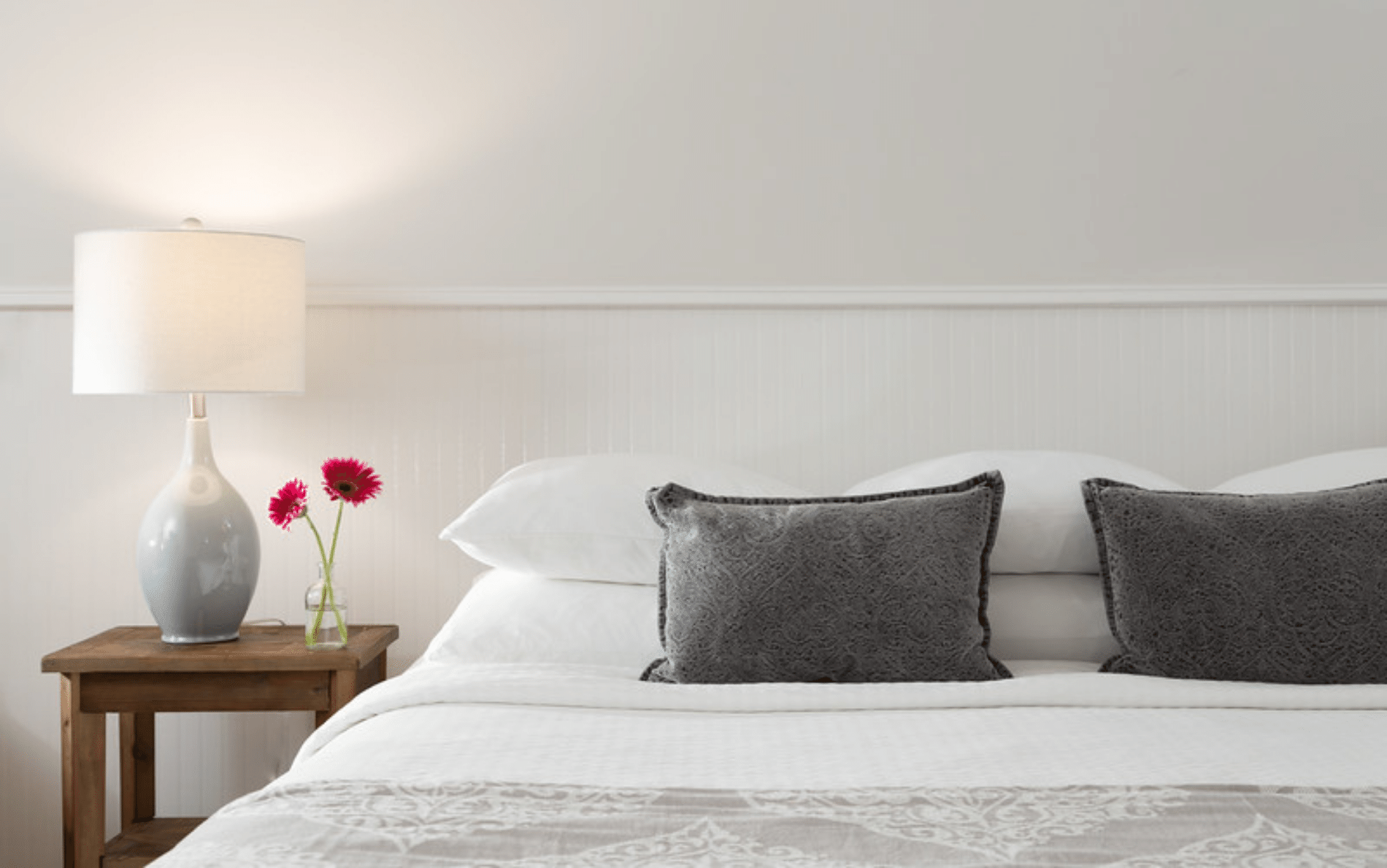 gray pillows on a white bed with pink flowers on a nightstand with a white light. 