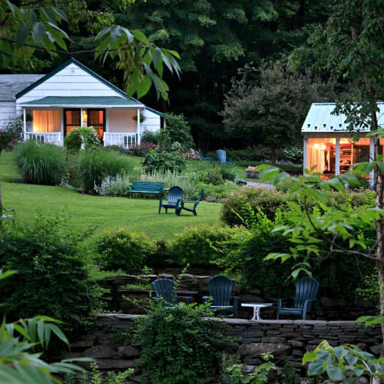 Sunken patio with stone walls and Adirondack chairs surrounded by grassy lawn with lush greenery and white cottage in background