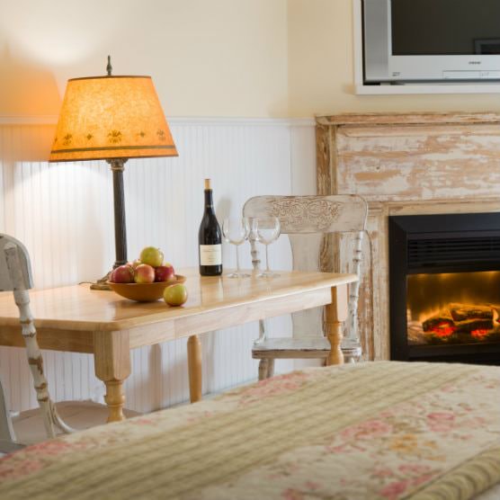 Two chairs and rectangular wood table topped with lamp, wine bottle and glasses, bowl of fruit with fireplace in background