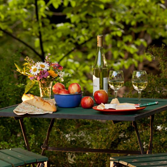 Slatted, outdoor table topped with wine bottle and glasses, apples in blue bowl, French bread, and vase of fresh wildflowers