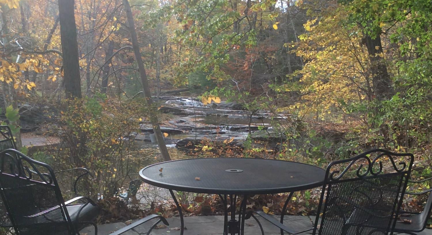 Black round metal table and chairs on a patio overlooking a stream and the woods with trees changing colors