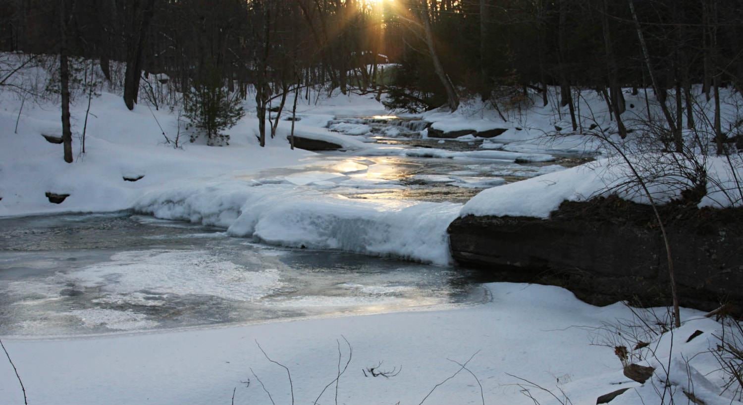 White snow and ice covered stream and woods with a golden setting sun in the distance