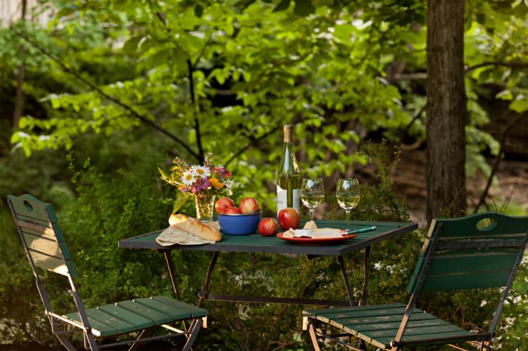 Green table and chairs topped with fresh flowers and apples, French bread, white wine, surrounded by lush greenery