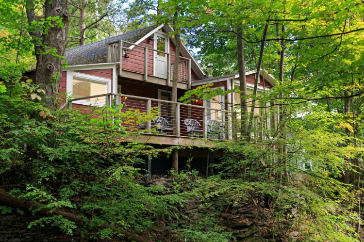 Red sided cottage with white trim, upper and lower decks, surrounded by woods with green leafed trees