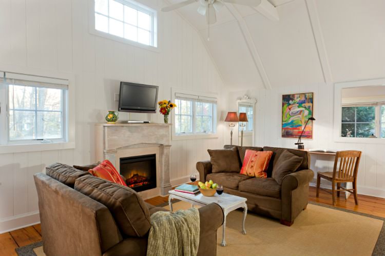 White vaulted room with wood floors, several windows, tv over fireplace, two upholstered love seats and writing desk with chair