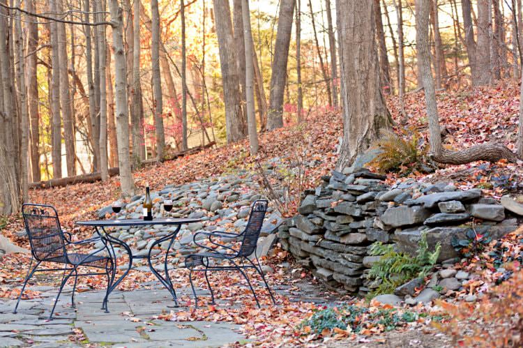 Black table and two chairs on a patio surrounded by woods, almost bare trees, and fallen leaves on the ground