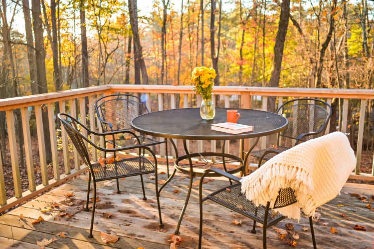 Wood deck with black metal table and chairs topped with fresh flowers, mug, book, and throw, overlooking woods
