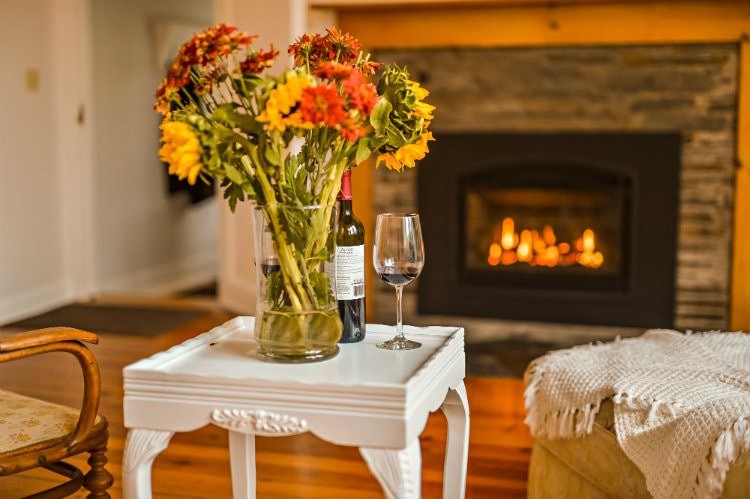 Close up of small white end table topped with fresh flowers and wine, and view of lit fireplace in background