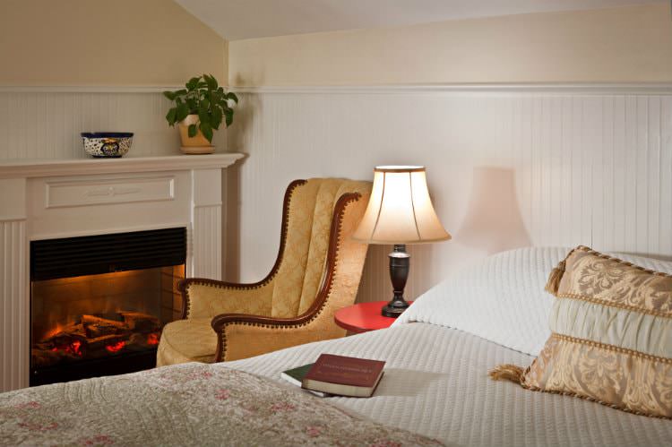 White covered bed with floral quilt, nightstand with lit lamp, and upholstered chair near a glowing fireplace