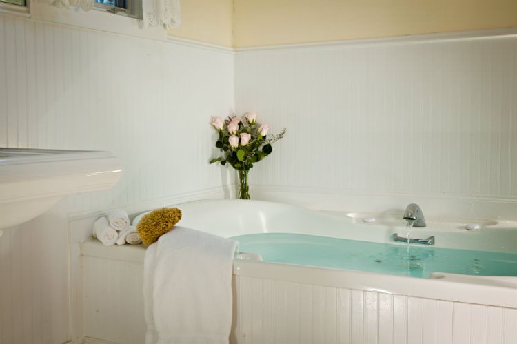 Cream bathroom with white beadboard, pedestal sink and soaker tub full of water with white towels and fresh flowers nearby