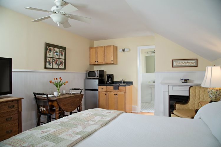 Beige and white vaulted room with fireplace, sitting chair, bed, drop-leaf table and chairs, kitchenette, and flat screen tv