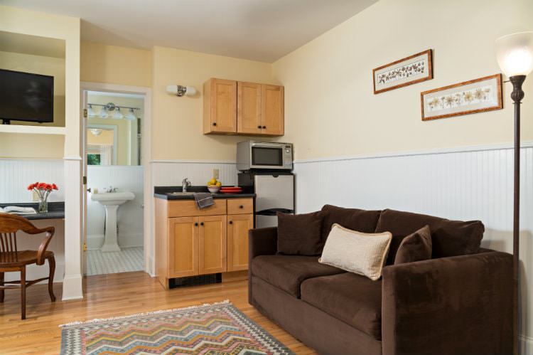 Beige room with white beadboard, kitchenette, brown upholstered couch, desk and chair and flat screen tv