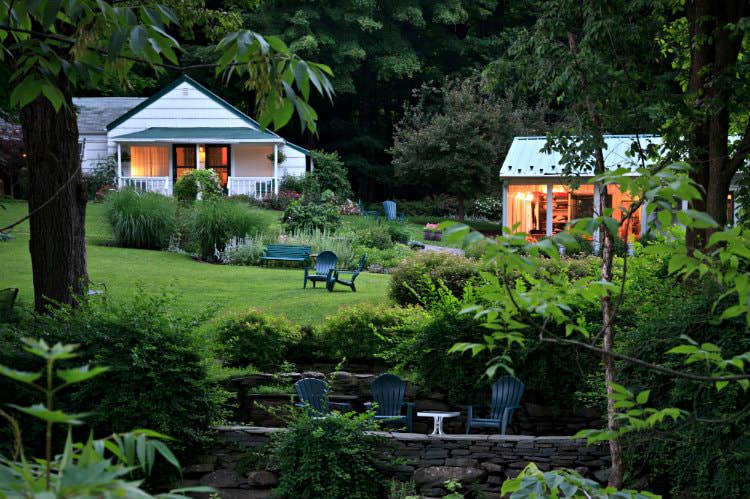 Sunken patio with stone walls and Adirondack chairs surrounded by grassy lawn with lush greenery and white cottage in background