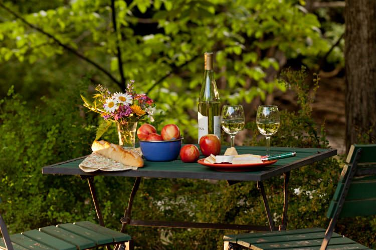 Green table topped with fresh flowers, white wine, red apples, French bread, surrounded by lush green woods
