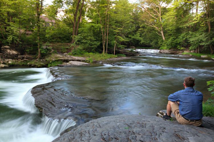 Man sitting on a rock ledge overlooking rippling stream with shallow waterfall surrounded by lush green woods