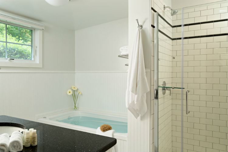 White bathroom with tiled walk-in shower with glass door, tub full of water, window, white towels and fresh flowers