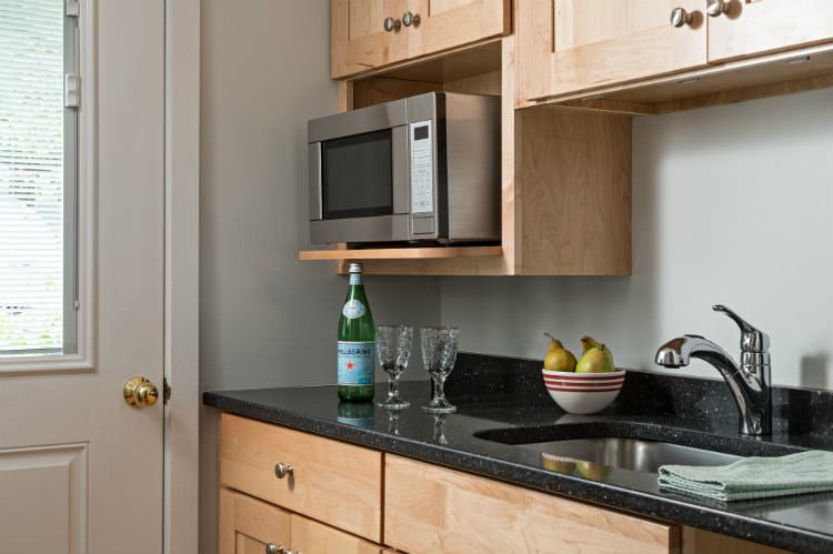 Close up of natural wood kitchen with dark stone tops, stainless steel microwave and half-glass door nearby