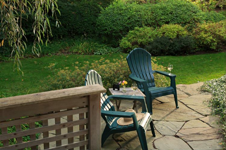 Two green Adirondack chairs with table on flagstone patio surrounded by lush green grass and shrubs