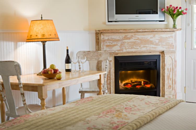 Beige and white room with fireplace, flat screen tv, wood table with white chairs topped with lamp, apples and wine