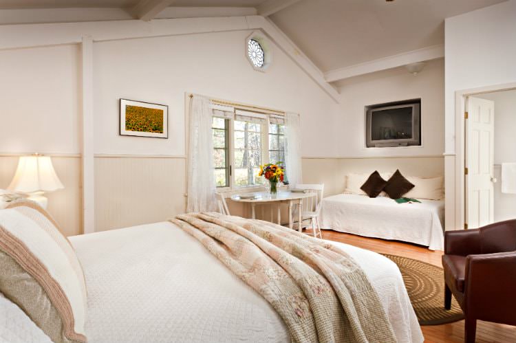 White vaulted room with beige beadboard, hardwood floor, bed with white and floral quilts, flat screen tv and dining table