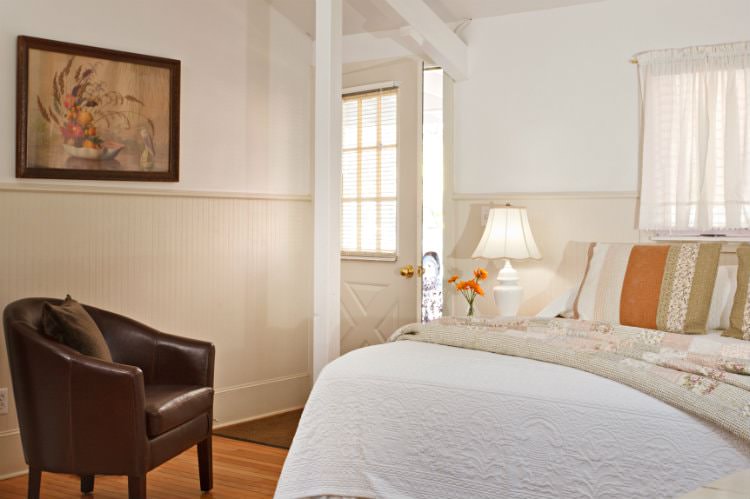 White room with beige beadboard, hardwood floor, bed with white and floral quilts, brown leather club chair, and natural light