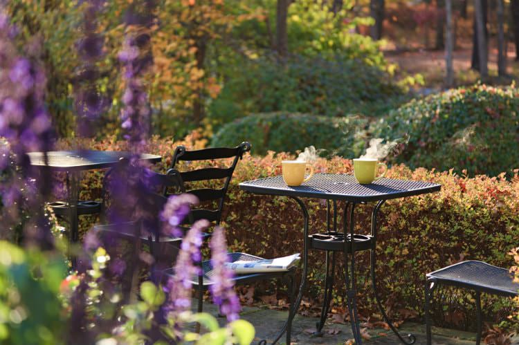 Black patio tables and chairs surrounded by shrubs, woods, and flowering plants