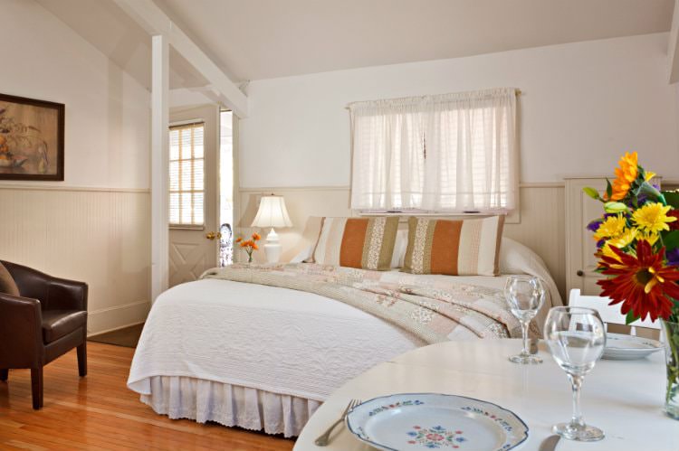 White room with beige beadboard, hardwood floor, bed with white and floral quilts, brown leather club chair and dining table