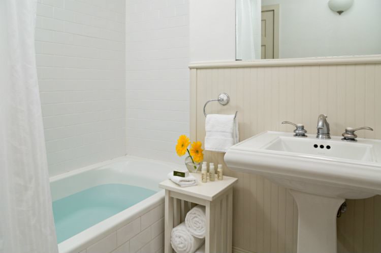 White bathroom with white tiled tub and shower, beige beadboard, white pedestal sink with mirror, and white towels