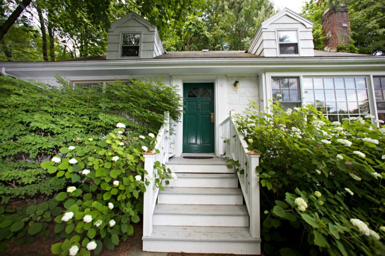 Exterior view of white cottage with green door, wood steps leading to the front door surrounded by green shrubs and trees