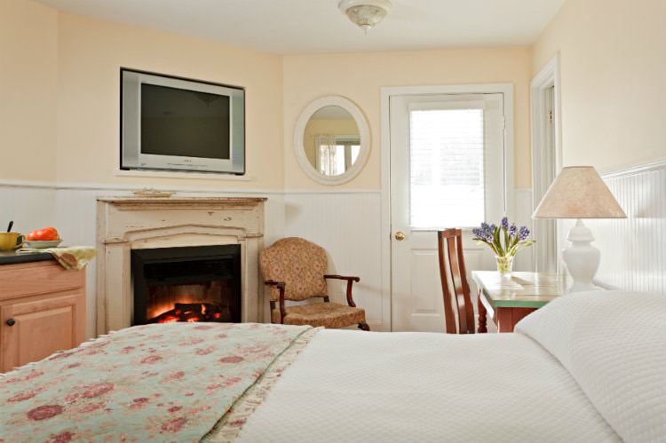 Beige room with white beadboard, bed with white and floral quilts, desk and chair, and fireplace with flat screen tv