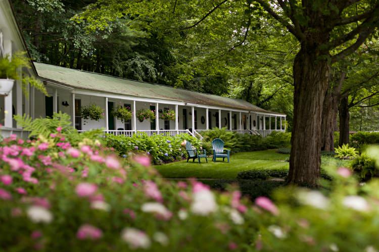 Two Adirondack chairs in grassy lawn surrounded by white inn, flowering shrubs and lush green trees