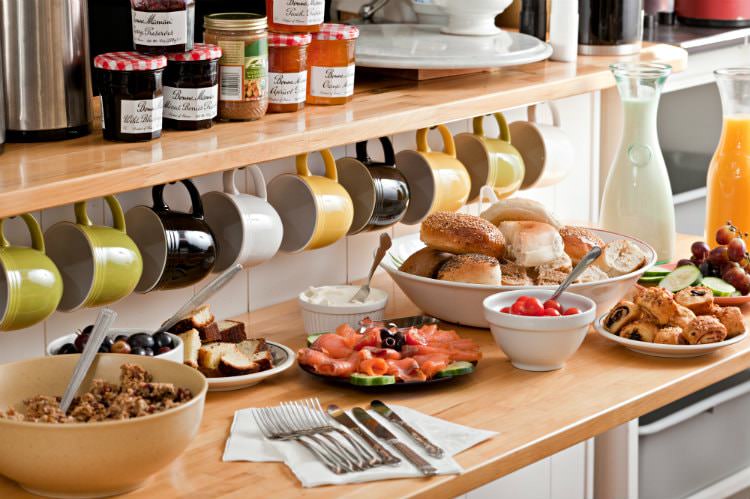 Butcher block counter topped with breakfast foods under a row of colorful mugs hanging from a wood shelf