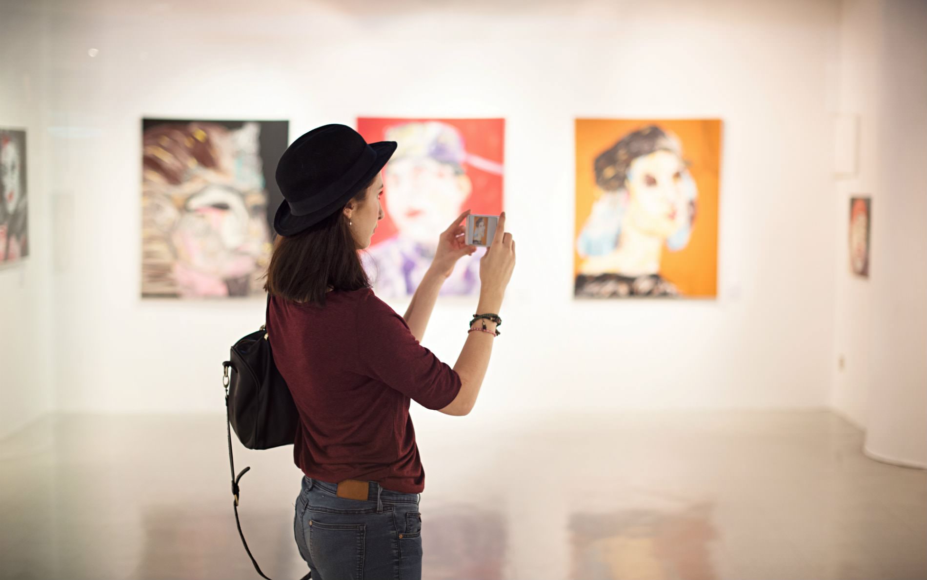 Brown haired woman with black hat, red shirt, and jeans taking a photograph of art hanging on white walls