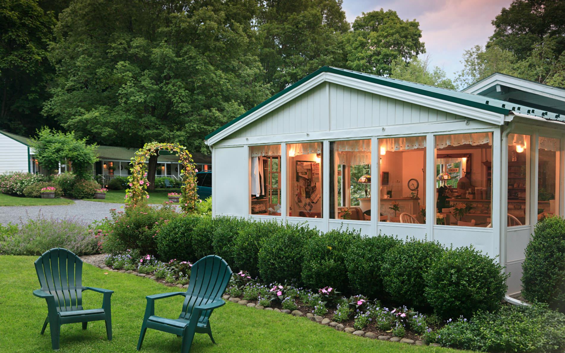 White cottage with lots of windows surrounded by a grassy lawn with two Adirondack chairs, green shrubs and flowers