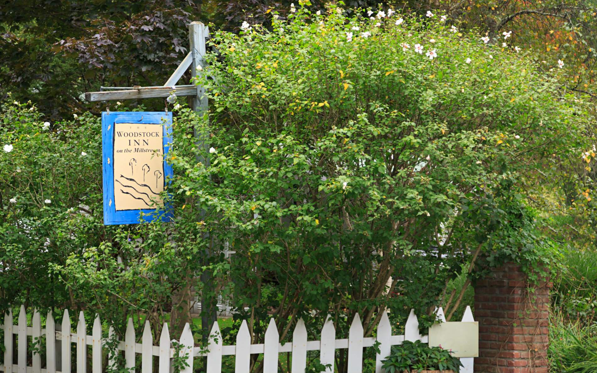 Beige and blue Woodstock Inn sign hanging from rustic wood post over a white picket fence surrounded by greenery