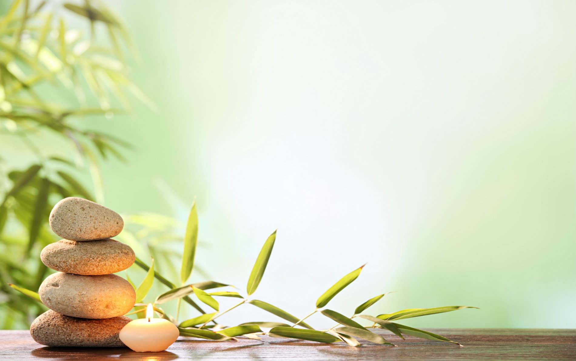 Four stacked rocks and small creamy lit candle on a wood table near a green plant, surrounded by diffused light